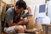 Full body of concentrated male master in apron sitting at table while sculpting with brown clay on throwing wheel — Stock Photo