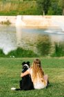 Vue arrière d'une adolescente méconnaissable embrassant un chien de Border Collie moelleux assis sur une pelouse près d'un étang dans un parc d'été — Photo de stock