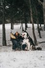 Junge ethnische Dame in Oberbekleidung umarmt niedlichen Husky-Hund, während sie im Winter in verschneiten Wäldern neben grünen Fichten hockt — Stockfoto