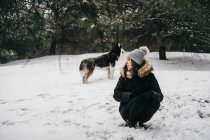 Young ethnic lady wearing outerwear with cute husky dog while crouching in snowy woods near green spruces in winter day — Stock Photo