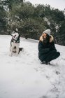 Jovem senhora étnica vestindo outerwear com cão bonito husky enquanto se agacha em florestas nevadas perto de abetos verdes no dia de inverno — Fotografia de Stock