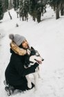 Молода етнічна леді, одягнена в верхній одяг, обіймає милого собаку-чоловіка, прив'язуючись у засніженому лісі біля зелених ялин в зимовий день — стокове фото