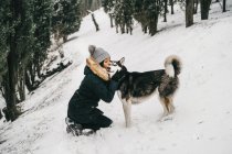 Jeune femme ethnique souriante portant des vêtements de plein air étreignant et embrassant chien husky mignon tout accroupi dans les bois enneigés près des épicéas verts dans la journée d'hiver — Photo de stock