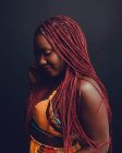 Mulher afro-americana alegre com tranças cor-de-rosa em pé sobre fundo preto em estúdio e olhando para baixo — Fotografia de Stock