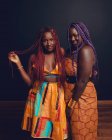 Conteúdo Amigos do sexo feminino afro-americanos com tranças coloridas e roupas laranja brilhante em pé no fundo escuro em estúdio e olhando para a câmera — Fotografia de Stock