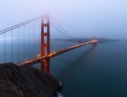 Pont Golden gate avec des lumières éclatantes sur la mer calme contre la ville côtière couverte de brouillard le soir à San Francisco — Photo de stock