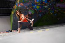 Longitud completa de la joven escaladora enfocada en ropa deportiva estirando las piernas mientras hace ejercicio en el centro de bouldering - foto de stock