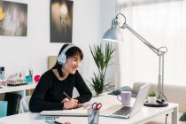 Усміхнена жінка-художниця малює в ескізі, сидячи за столом у навушниках і переглядаючи онлайн-урок на нетбуці вдома — стокове фото