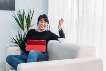 Glad female sitting on sofa and watching funny movie on tablet while relaxing at weekend at home — Stock Photo