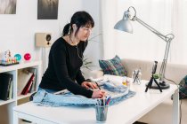 Vista lateral de la experta diseñadora femenina escuchando música en auriculares y dibujando boceto en chaqueta de mezclilla en la mesa en casa - foto de stock