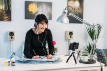Vista lateral de designer feminina qualificada ouvindo música em fones de ouvido e desenho de jaqueta de ganga na mesa em casa — Fotografia de Stock
