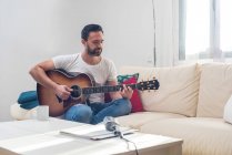 Урожай бородатого чоловіка-музиканта, який грає на акустичній гітарі на дивані біля старовинного мікрофона, розміщеного на столі — стокове фото