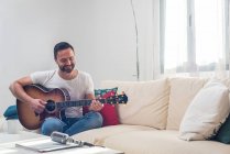 Ein bärtiger Musiker spielt Akustikgitarre auf einem Sofa in der Nähe eines Mikrofons, das auf dem Tisch steht — Stockfoto