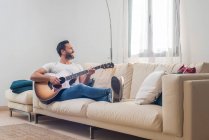 Corpo inteiro de feliz adulto músico masculino étnico em roupas casuais relaxando no sofá confortável e tocando guitarra acústica em casa — Fotografia de Stock