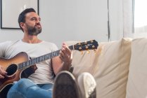 Все тело взрослого этнического мужчины-музыканта в повседневной одежде расслабляется на удобном диване и играет на акустической гитаре дома — стоковое фото