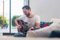 Crop of bearded male musician playing acoustic guitar on sofa near window at home — Stock Photo