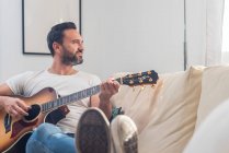 Voller Körper eines erwachsenen ethnischen männlichen Musikers in lässiger Kleidung, der es sich auf einem bequemen Sofa bequem macht und zu Hause Akustikgitarre spielt — Stockfoto