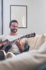 Счастливый взрослый этнический мужчина музыкант в повседневной одежде расслабляется на удобном диване и играет на акустической гитаре дома — стоковое фото