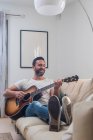 Повне тіло щасливого дорослого етнічного чоловіка-музиканта в повсякденному одязі, що розслабляється на зручному дивані і грає на акустичній гітарі вдома — стокове фото