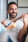 Feliz adulto étnico masculino músico em roupas casuais relaxando no sofá confortável e tocando guitarra acústica em casa — Fotografia de Stock
