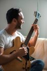 Musicista adulto di etnia maschile in abiti casual che si rilassano sul comodo divano e suonano la chitarra acustica a casa — Foto stock