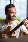 Счастливый взрослый этнический мужчина музыкант в повседневной одежде расслабляется на удобном диване и играет на акустической гитаре дома — стоковое фото