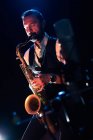 Side view of concentrated bearded male musician in elegant outfit playing saxophone while standing on stage during concert — Stock Photo