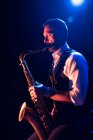 Бородатый музыкант в элегантном наряде, играющий на саксофоне, стоя на сцене во время концерта — стоковое фото