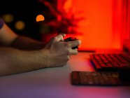 Кадрирование неузнаваемый мужчина геймер с помощью геймпада и играть в видеоигры, сидя за столом с компьютером в темной комнате — стоковое фото