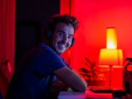 Вид сбоку красивый счастливый мужчина в повседневной рубашке и беспроводных наушниках опираясь на стол с компьютером и глядя на камеру с улыбкой в темной комнате — стоковое фото