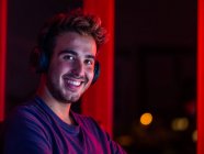Handsome happy male wearing casual shirt and wireless headphones leaning on desk with computer and looking at camera with smile in dark room — Stock Photo