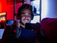 Zufriedener junger Mann in Freizeitkleidung und kabellosen Kopfhörern surft im Handy, während er sich im dunklen Raum auf dem Sofa ausruht — Stockfoto