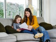 Zufriedene weibliche Teenager, die die Lehrbuchseite betrachten, während sie Zeit mit Geschwistern auf der Couch zu Hause verbringen — Stockfoto