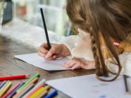 Обрізати анонімну маленьку дівчинку, малюючи різнокольоровими олівцями на паперовому аркуші у світлій кімнаті — стокове фото