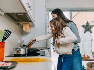 Весела молода мати піднімає милу усміхнену дочку, перемішуючи їжу на сковороді, готуючи разом на сучасній кухні — стокове фото