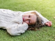 Adorable niña alegre vistiendo suéter blanco acogedor acostado en el prado herboso y mirando a la cámara con sonrisa - foto de stock