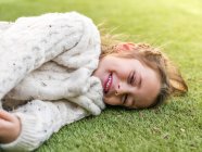 Entzückend fröhliches kleines Mädchen im kuscheligen weißen Pullover, das auf der Wiese liegt und lächelnd in die Kamera schaut — Stockfoto