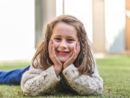 Adorable niña alegre vistiendo suéter blanco acogedor acostado en el prado herboso y mirando a la cámara con sonrisa - foto de stock