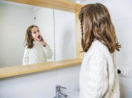 Вид збоку щаслива маленька дівчинка в затишному білому светрі дивиться в дзеркало з зубною посмішкою, стоячи з зубною щіткою в сучасній ванній — стокове фото