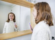 Счастливая маленькая девочка в уютном белом свитере смотрит в зеркало с зубной улыбкой, стоя с зубной щеткой в современной ванной комнате — стоковое фото