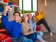 Lächelnde Studentinnen sitzen mit Notizbuch auf dem Sofa und machen in der Pause Selbstporträts mit dem Handy — Stockfoto