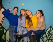 Mujeres jóvenes felices sonriendo brillantemente mientras están de pie juntos contra la pared pintada de colores y mirando a la cámara - foto de stock