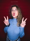 Young female doing grimace showing peace gesture while standing against red striped background — Stock Photo