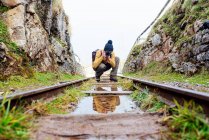 Full body young female photographer in warm sweater and hat sitting on haunches on railroad between rough rocks and taking a picture — Stock Photo