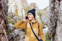 Positivo giovane femmina in abbigliamento casual e cappello rispondere telefonata mentre in piedi in mezzo a formazioni rocciose e guardando lontano contento — Foto stock
