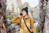 Mujer joven positiva en ropa casual y sombrero respondiendo a una llamada telefónica mientras está de pie en medio de formaciones rocosas y mirando hacia otro lado contenta - foto de stock