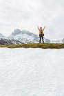 Все тело веселой женщины в теплой одежде, поднимающей руки в волнении, стоя на огромной горной долине, покрытой снегом и окруженной скалистыми суровыми горами — стоковое фото