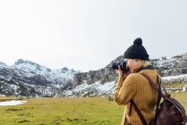 Visão lateral focou jovem fotógrafa em suéter quente tirando fotos na câmera fotográfica profissional de majestosas montanhas ásperas no planalto gramado no dia de outono claro — Fotografia de Stock