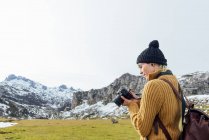 Side view focused young female photographer in warm sweater taking pictures on professional photo camera of majestic rough mountains on grassy highland on clear autumn day — Stock Photo