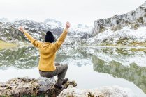 Анонимная женщина в тёплом свитере и черной шляпе сидит с руками, вытянутыми на грубом камне на спокойном озере, любуясь захватывающим пейзажем снежных скалистых гор. — стоковое фото